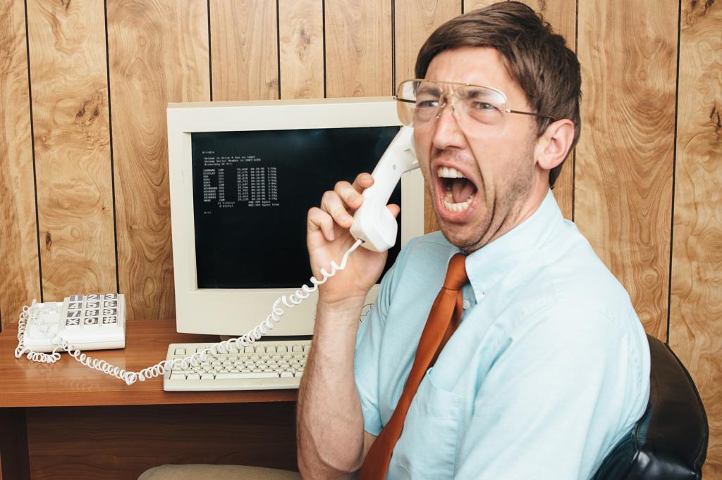 A man and office in 1980's - 1990's style, complete with vintage computer and technology of the time, yells in anger and frustration on the phone to tech support trying to figure out why his computer is broken. Wood paneling on the wall in the background. Horizontal with copy space.