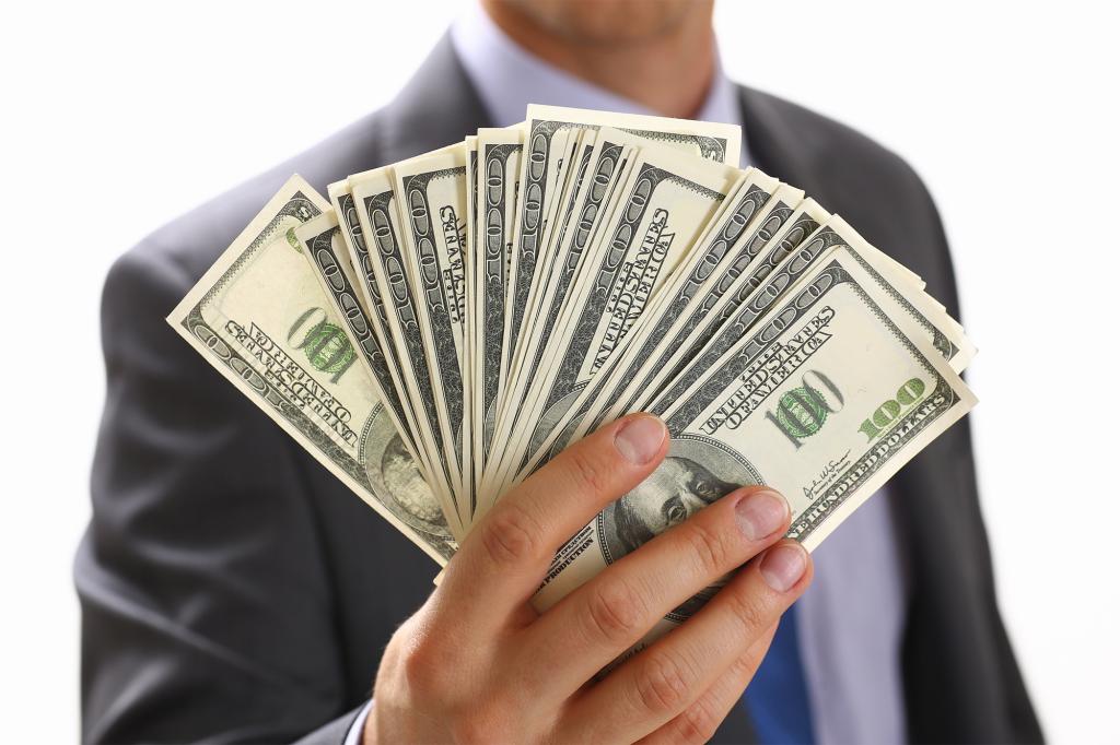 Man in suit and tie holding fanned out stack of hundred dollar bills.