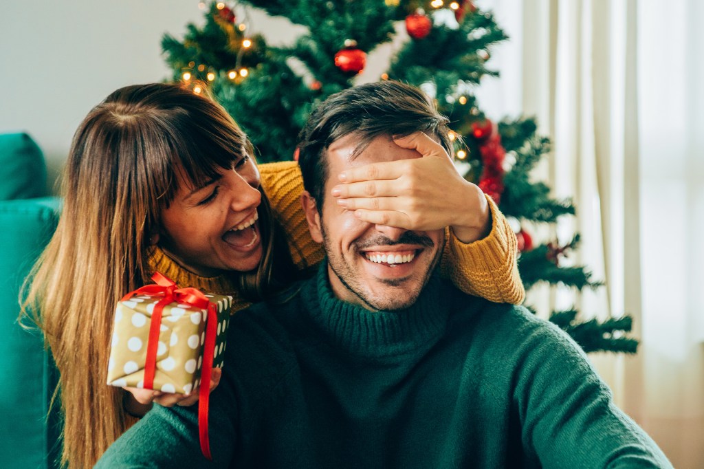 Beautiful young woman is standing behind her boyfriend with a gift in one hand and covered his eyes with the other hand.