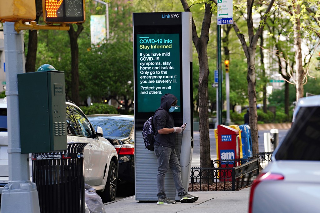 A man wearing a protective mask leans against a LinkNYC box.