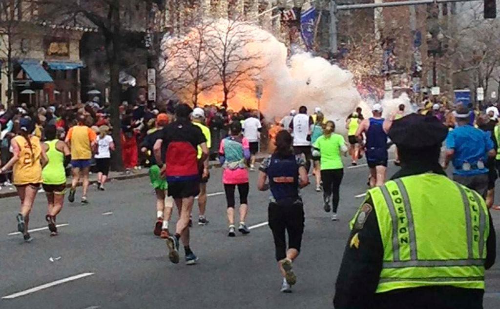 Runners continue to run towards the finish line of the Boston Marathon as an explosion erupts near the finish line of the race.