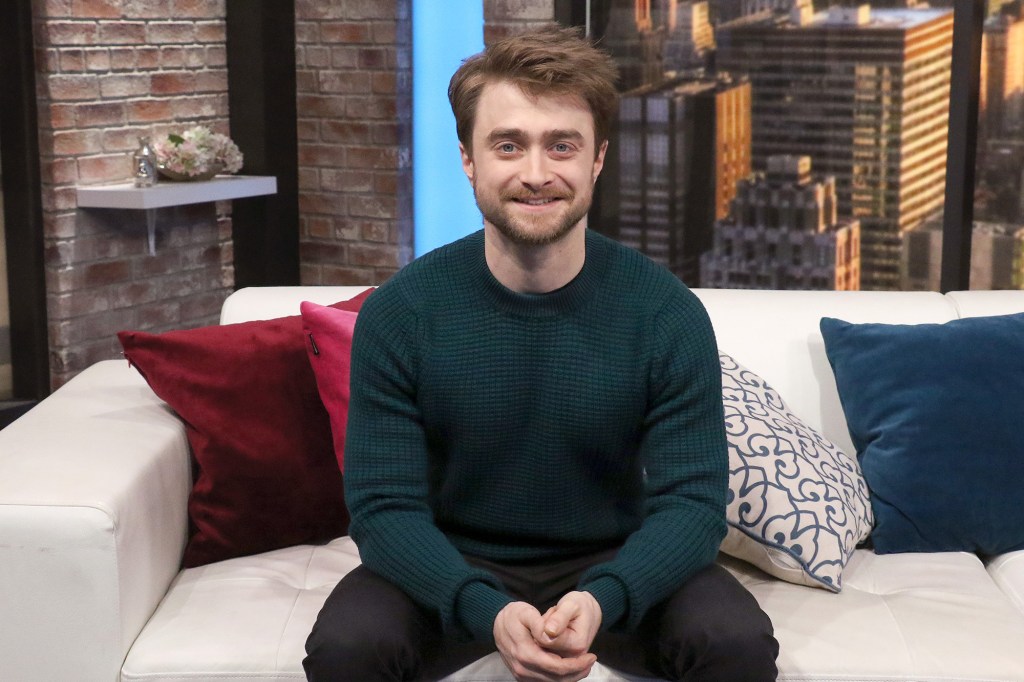 Daniel Radcliffe sits on a couch and smiles. 