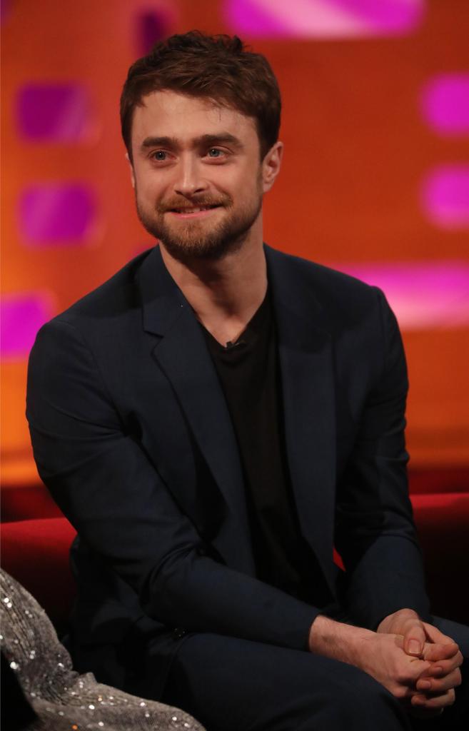 Daniel Radcliffe sits on a couch smiling.