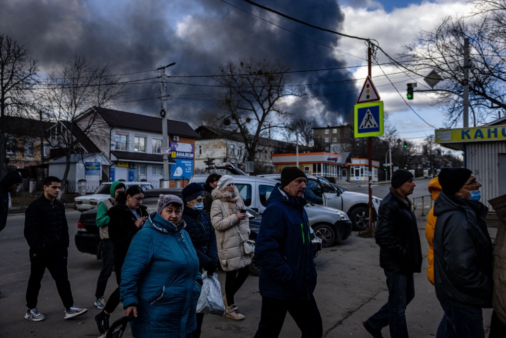 People stand in line in front of a supermarket while smoke billows over the town of Vasylkiv just outside Kiev.