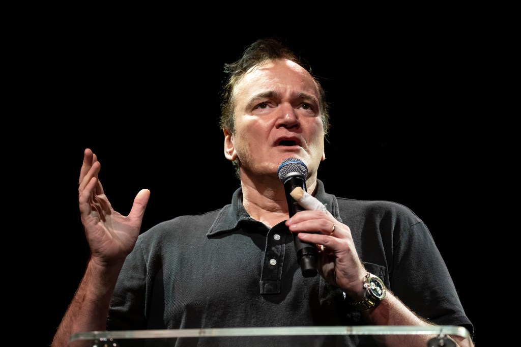 Quentin Tarantino holds a microphone and wears a black polo shirt