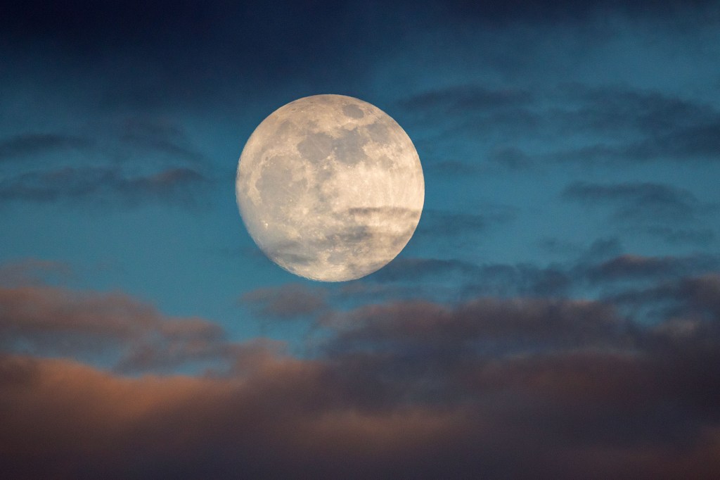 Photo of a full moon against a twilight sky with hazy clouds.