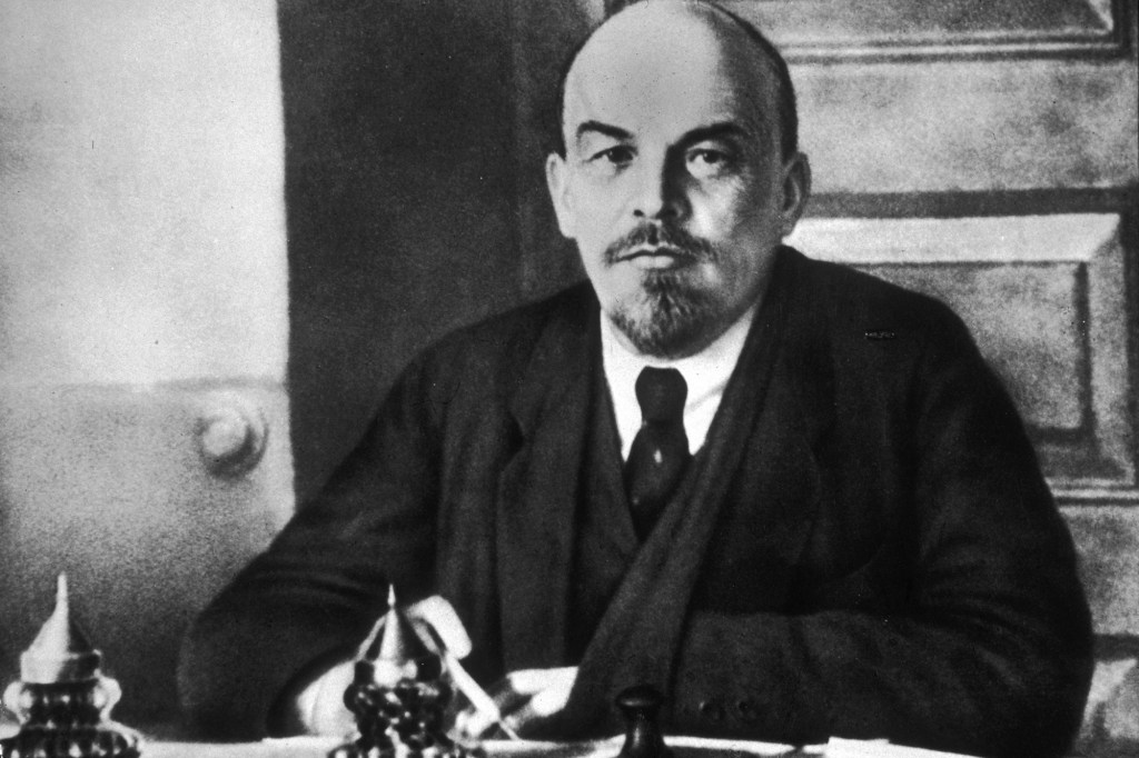 Black and white portrait of Vladimir Lenin in a suit seated at his desk.