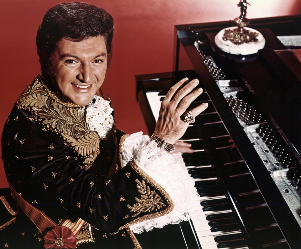 Portrait of Liberace playing his piano in an ornate jacket.