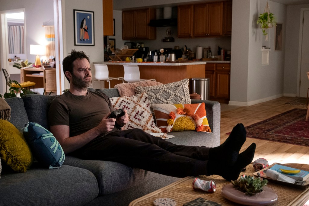 Bill Hader as hitman-turned-actor Barry Berkman in Season 3 of "Barry" on HBO. He's reclining on a couch with his shoes off while playing a video game.