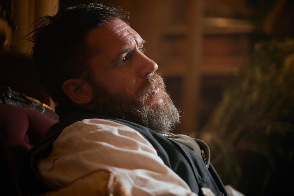 Tom Hardy from the side, sitting at a desk, looking serious with a beard.