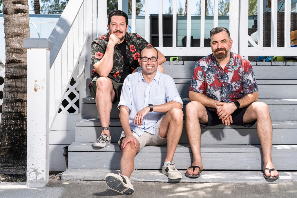 Photo of three of the "Impractical Jokers" sitting on the steps of a beach house. They're all wearing shorts and smiling.