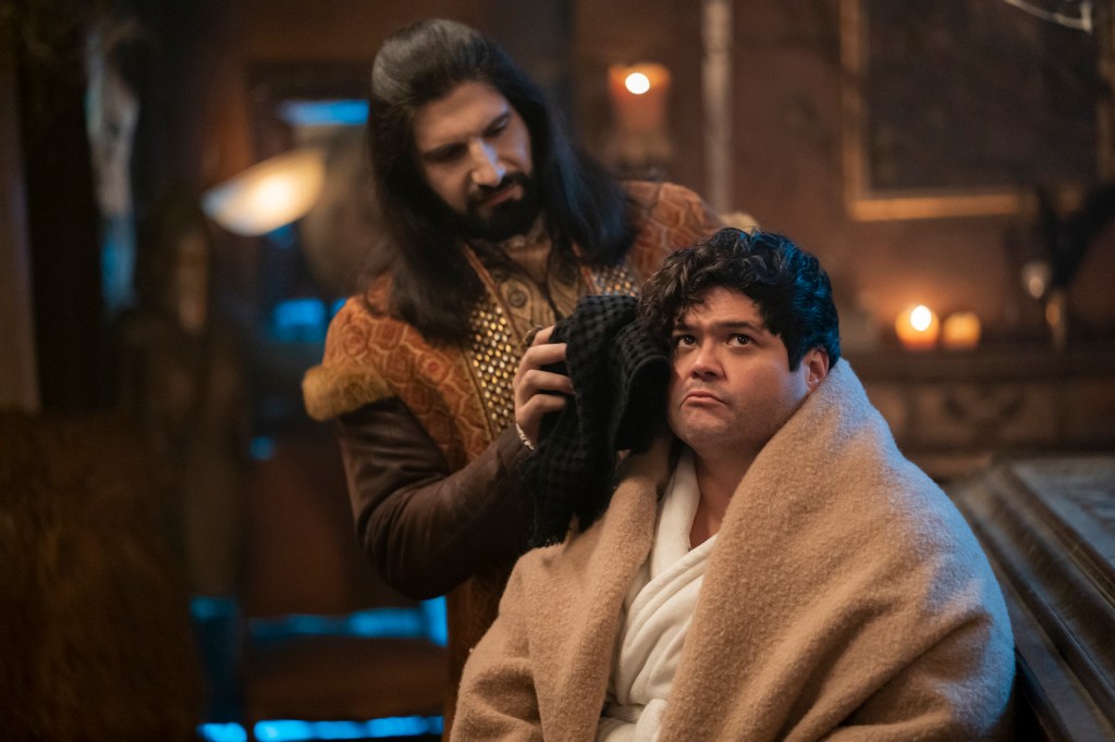 Photo of Kayvan Novak and Harvey Guillen as Nandor and Guillermo. Nandor is wiping Guillermo's face with a black rag. Guillermos has a blanket over him and is frowning.