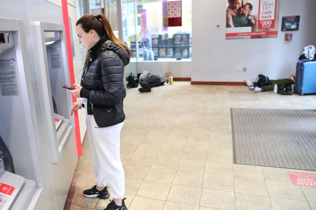 A woman withdrawing cash as two homeless people sleep in the bank lobby.