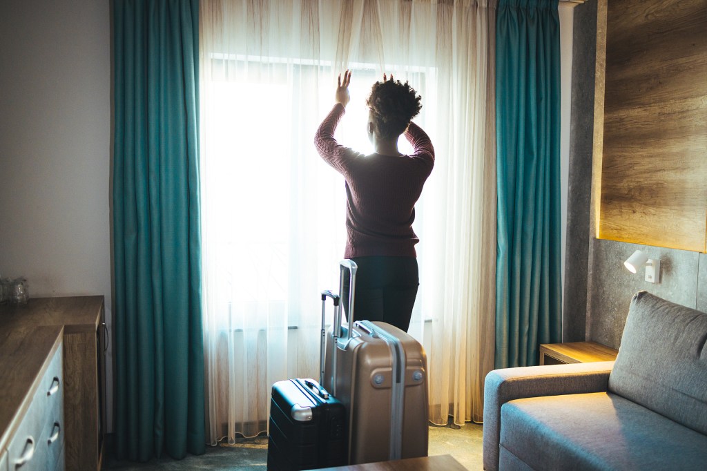 Luggage in modern hotel room with happy young adult female relaxing nearly window, African woman tourist looking to beautiful nature view. Time to travel, relaxation, journey, trip and vacation concepts