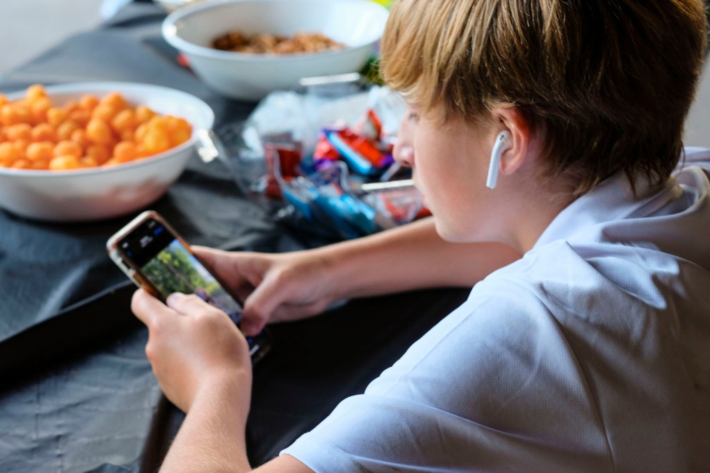 A preteen boy concentrates on his smartphone with ear buds and bowls of snacks.