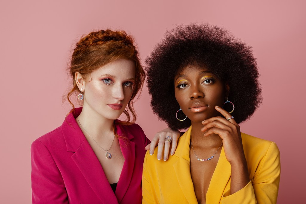 Red headed woman on left in hot pink jacket, woman on right with dark hair in yellow jacket, on pink background