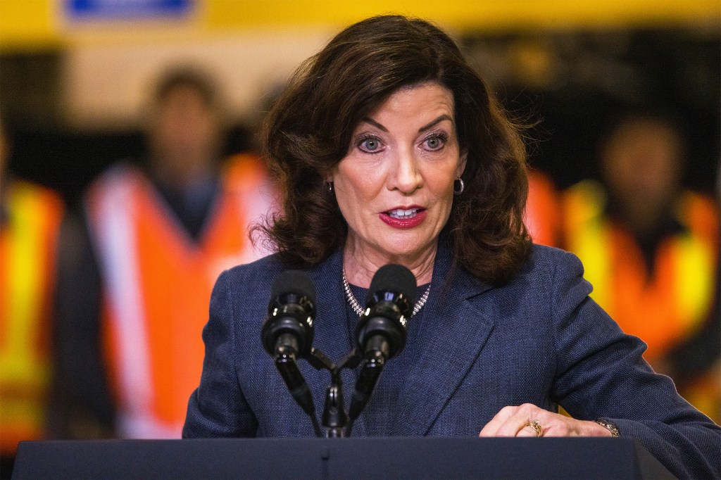 Hochul in a dark blue blazer looking at something from behind a podium with blurry people in orange safety vests in background