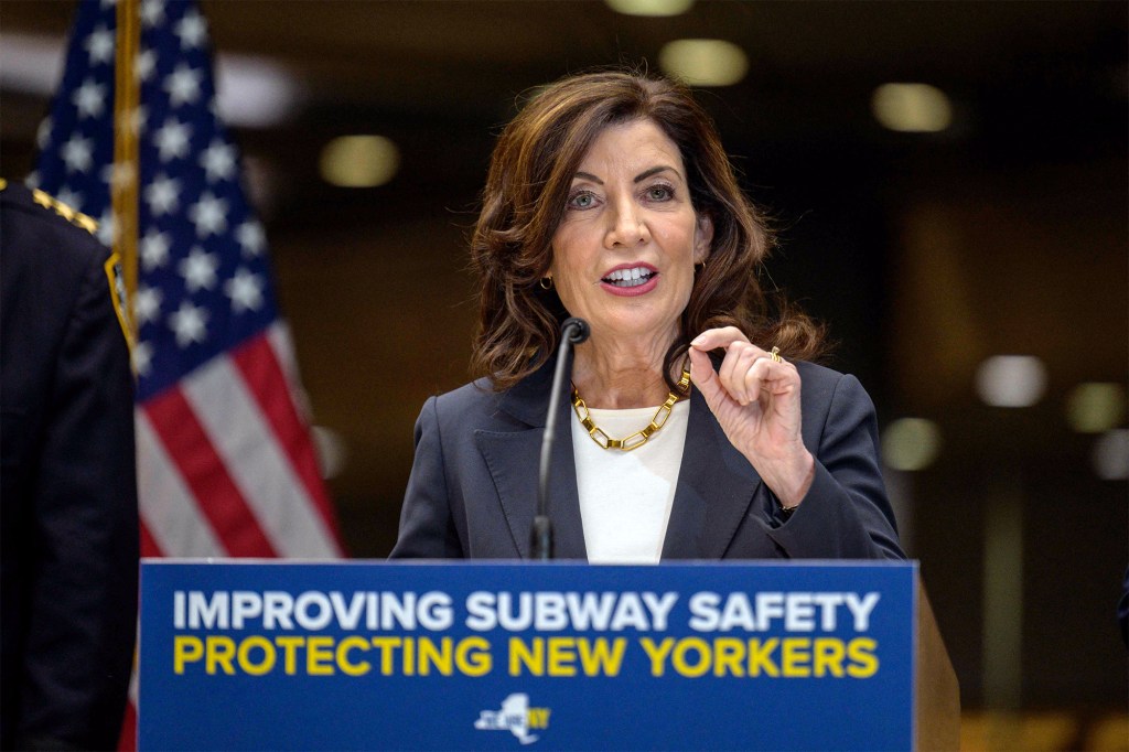 Hochul at a podium with thumb and index finger touching above a sign that says "Improving Subway safety protecting new yorkers"