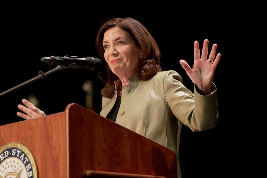 Gov. Kathy Hochul speaking at a podium with hands up in front of a black background