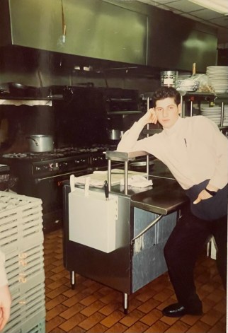 Photo of a young Steve Haxhiaj with his elbow on the counter of the restaurant's kitchen.