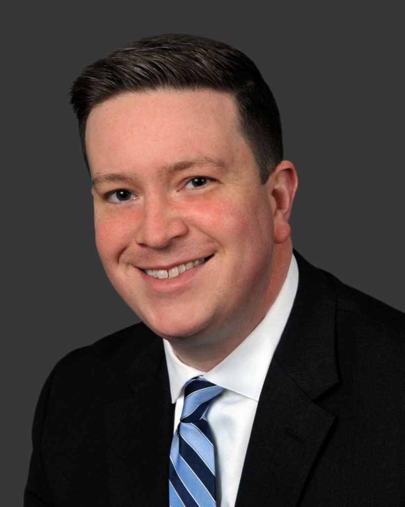 John McGowan headshot with a grew background. Black suit with a tie and a smile.