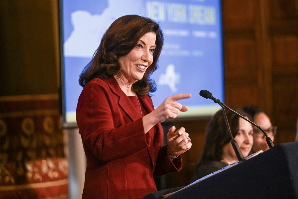 Hochul in a red outfit speaking in the red room within the State Capitol with staffers in background alongside a powerpoint budget presentation