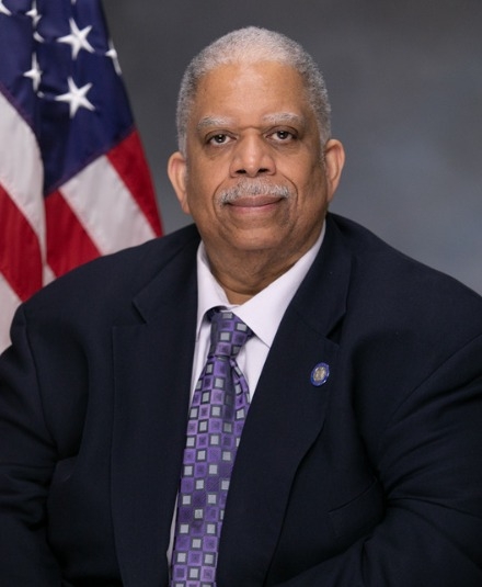 Leroy Comrie in a suit posing for the camera in front of an American flag and a grey background