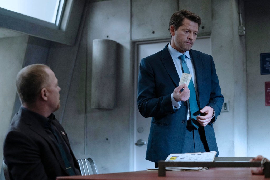 Misha Collins wears a suit and looks serious standing in an interrogation room. 