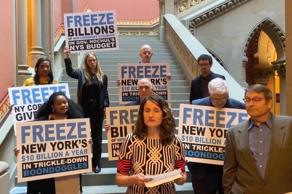 A woman standing on a staircase with people holding signs behind her criticizing economic development spending.