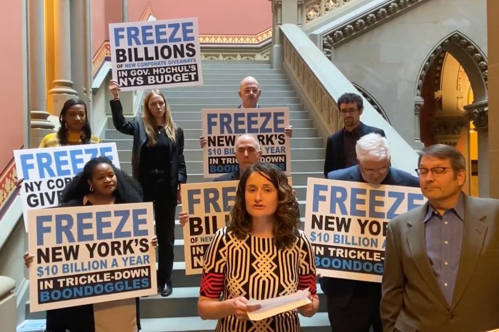 A woman standing on a staircase with people holding signs behind her criticizing economic development spending. 