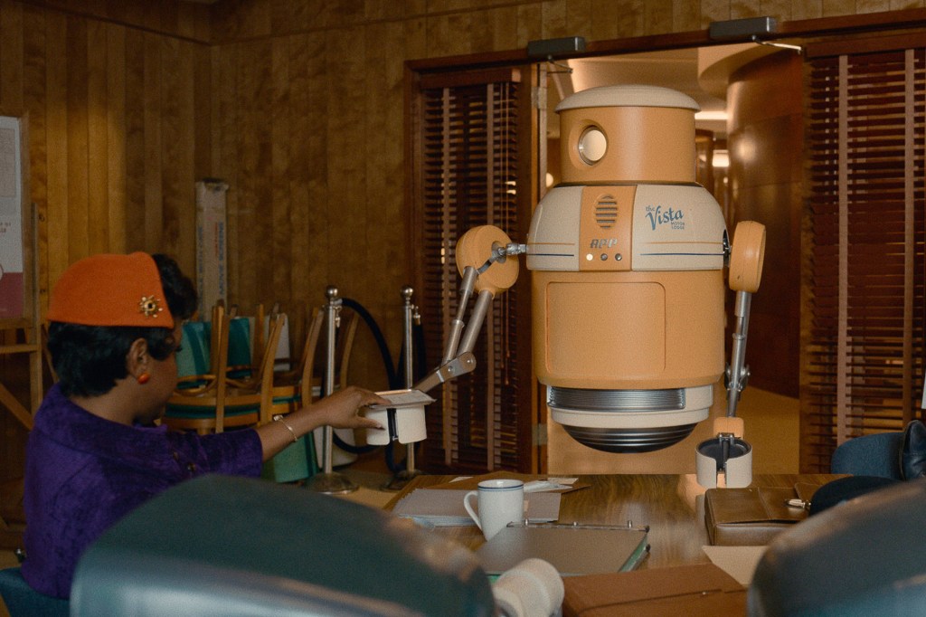 Scene from "Hello Tomorrow!" showing Shirley, played by Haneefah Wood, being assisted by a robot in her office, which is handing her what looks like a piece of paper with its metal arm.