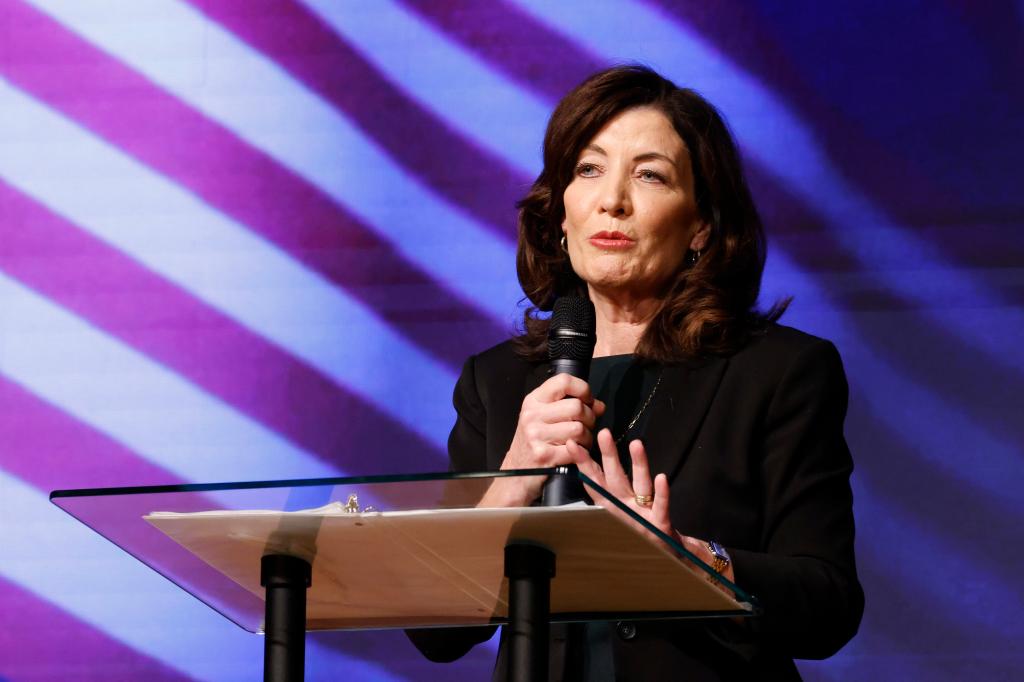 Kathy Hochul speaking with a microphone with a a projected background of red and white stripes.