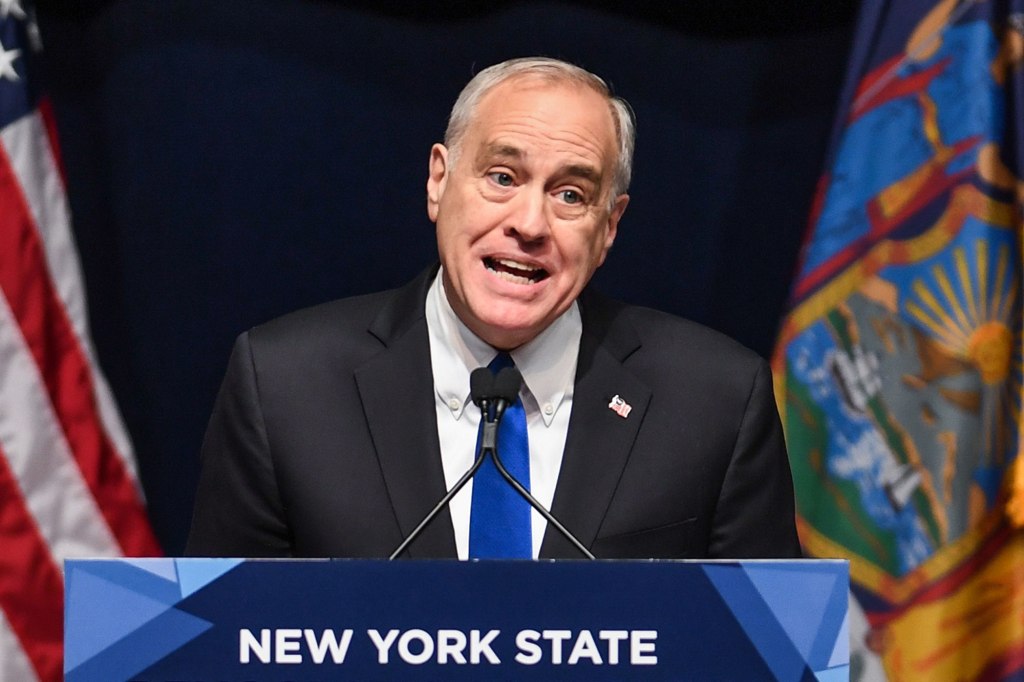 Thomas DiNapoli with a funny expression speaking at a podium in front of a dark background with flags