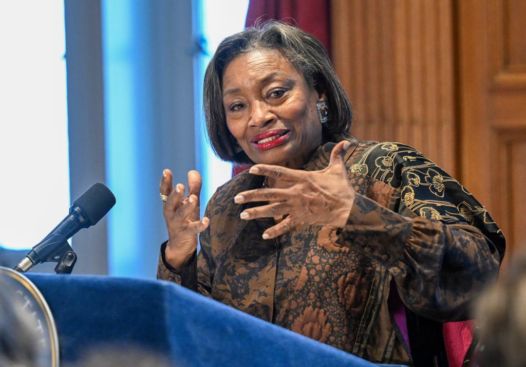 Andrea Stewart-Cousins speaking with gesticulating hands at a podium in front of a wooden wall