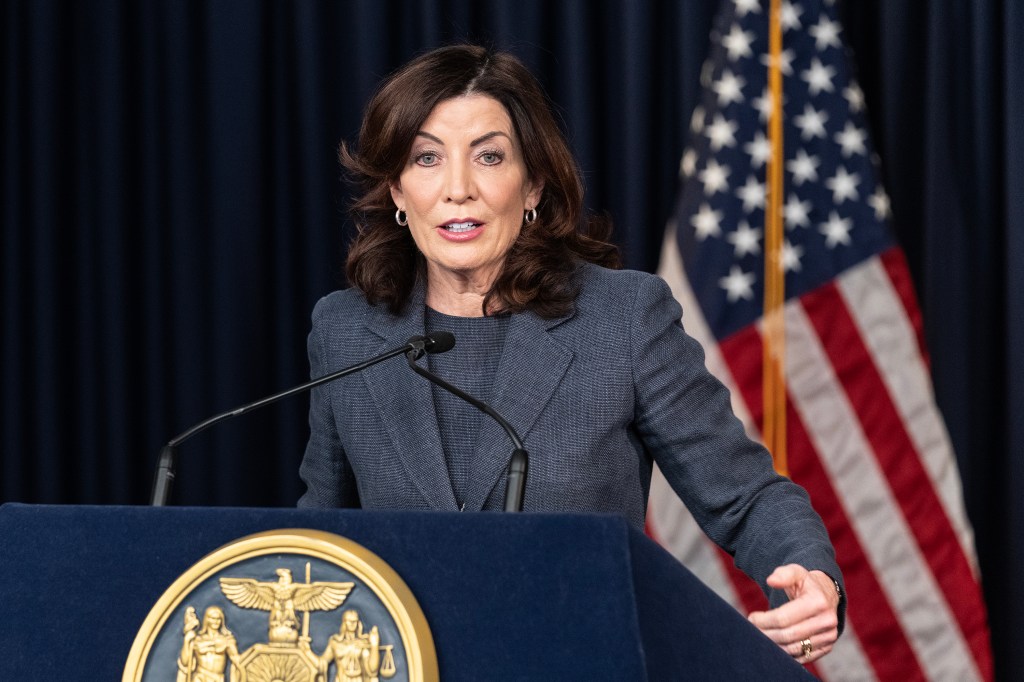 Kathy Hochul in a grey outfit speaking at a podium in front of a dark background and american flag