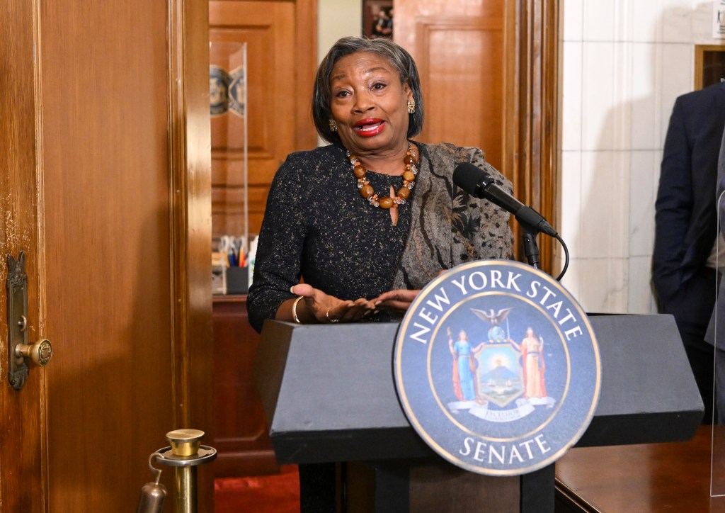 Andrea Stewart-Cousins with trademark scarf standing in a doorway at a podium with her hands held together upside down