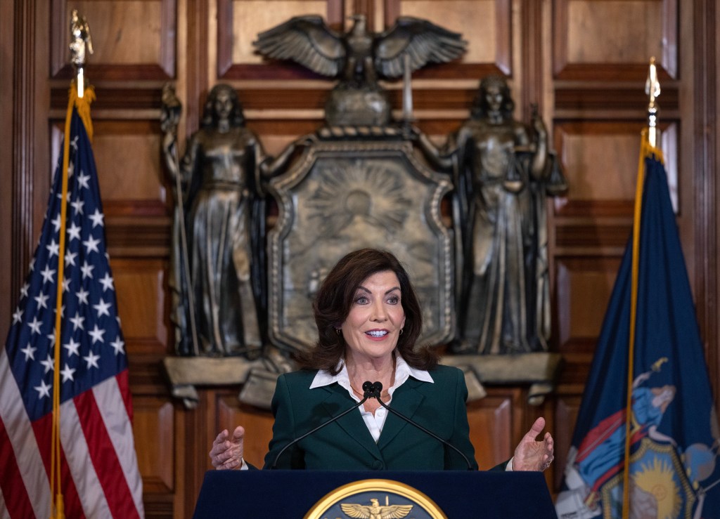 Kathy Hochul in a green jacket speaking in front a blurry state seal mounted on a wooden wall.