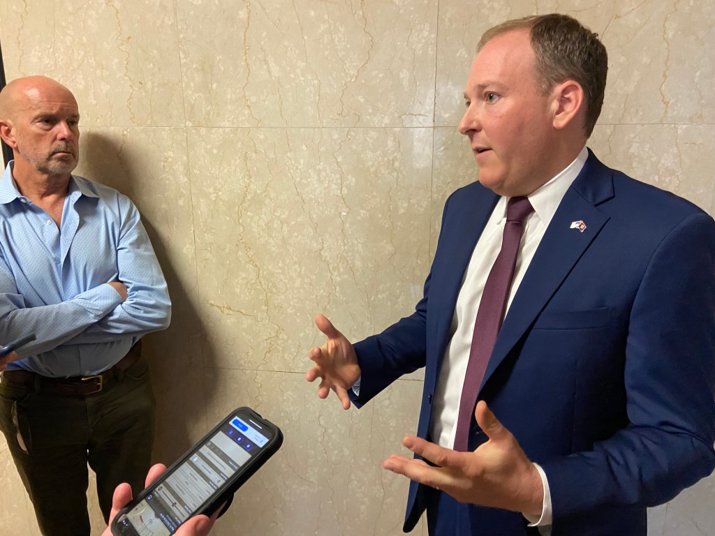 Lee Zeldin standing in front of a marble wall with gesticulating hands while a reporter looks at him critically. 