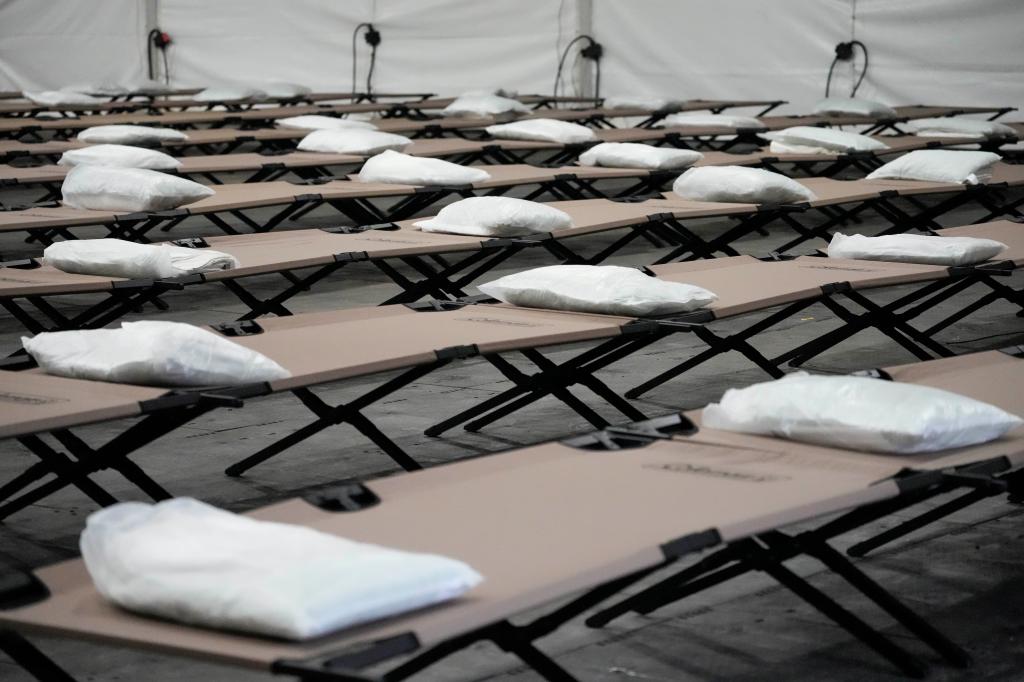 Bags containing a pillow, towel, and bed sheets are placed on cots inside the dormitory tent.