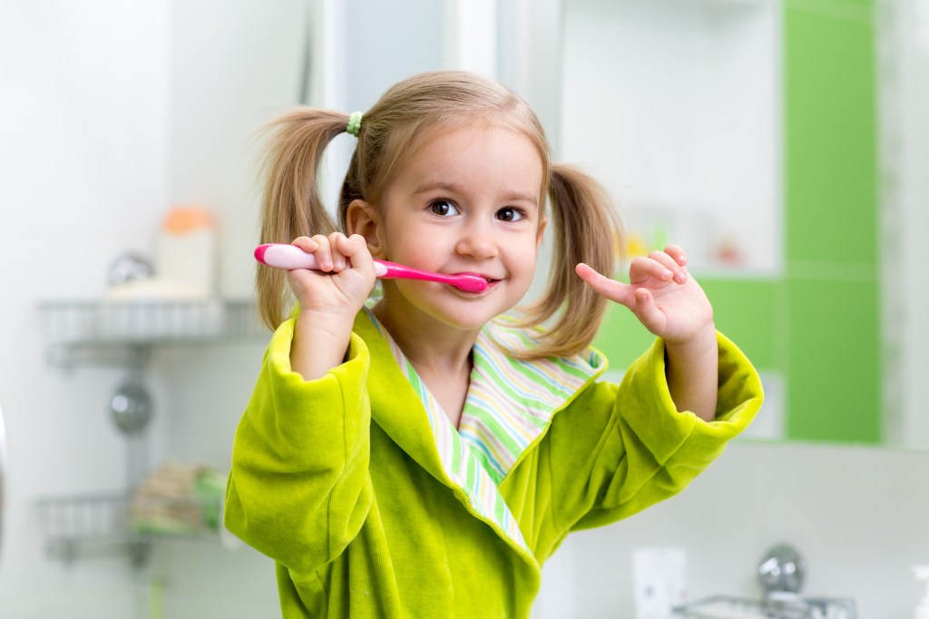 Photo of a child in pigtails brushing their teeth wearing a green robe. 