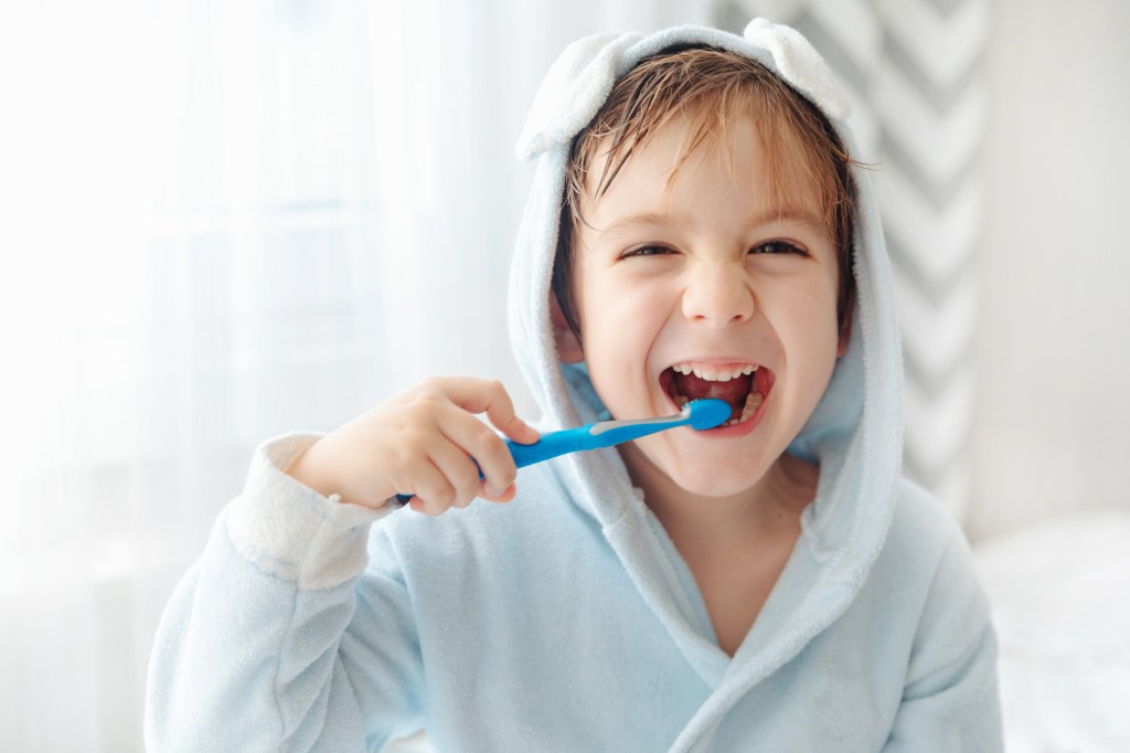 Photo of a kid brushing their teeth wearing a blue bathrobe with a hood attached. 