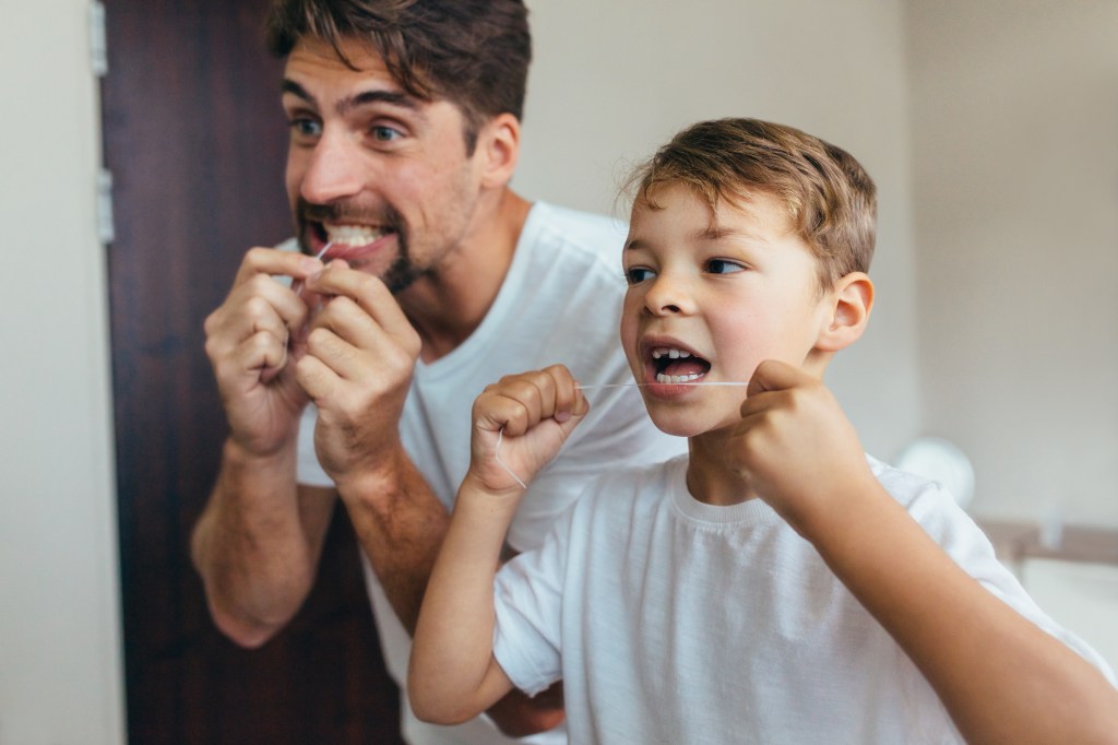Photo of a dad and son flossing their teeth together, both wearing white t-shirts. 