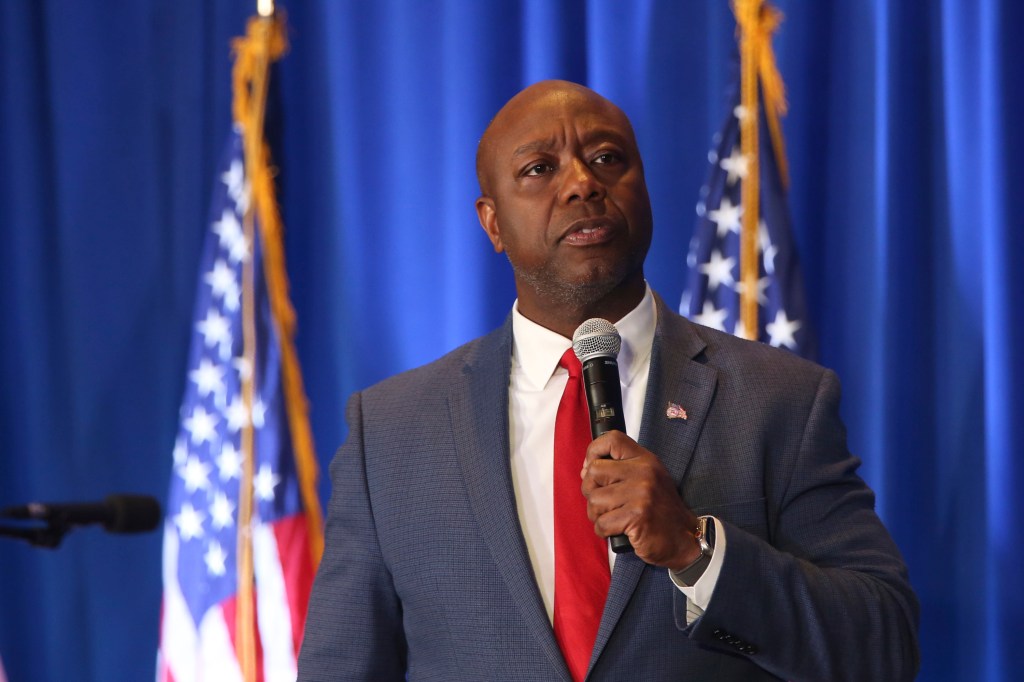 Tim Scott speaks into a microphone in front of American flags. 