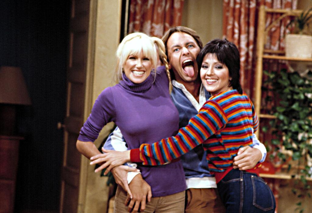 Suzanne Somers, John Ritter and Joyce DeWitt joking around on the set of "Three's Company" during its first season. Ritter's tongue is sticking out and he has he arms around Somers and DeWitt who are smiling broadly.