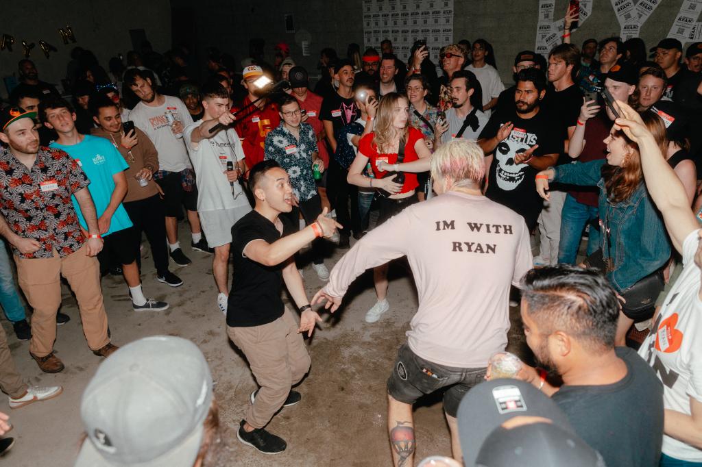 A crowded dancing in a warehouse setting wearing "Ryan" shirts.