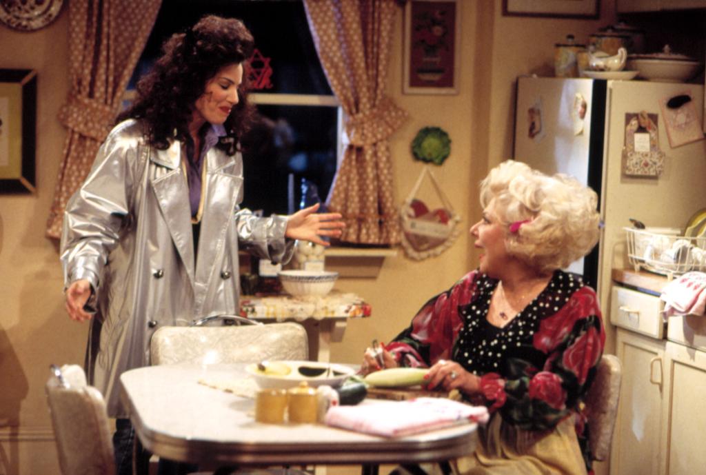 Fran Drescher and Renee Taylor as Fran and Sylvia in an episode of "The Nanny" on CBS. Renee is seated at the kitchen table and Fran is standing over her wearing a silver coat.