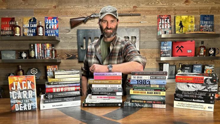 Jack Carr surrounded by books with a gun behind his head.