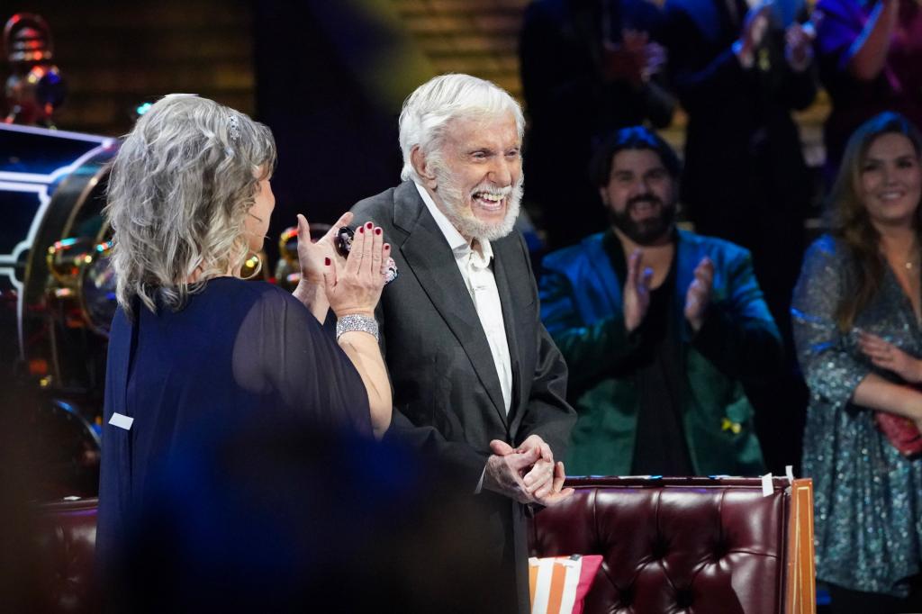 Dick Van Dyke, with his wife, Arlene Silver, walks onto the re-created set of "The Dick Van Dyke Show" with his wife, Arlene Silver. He's got a huge grin on his face, which is framed by a white beard. He's wearing a grey suit jacket and a white shirt. Arlene is wearing a blue dress and her back is to the camera.