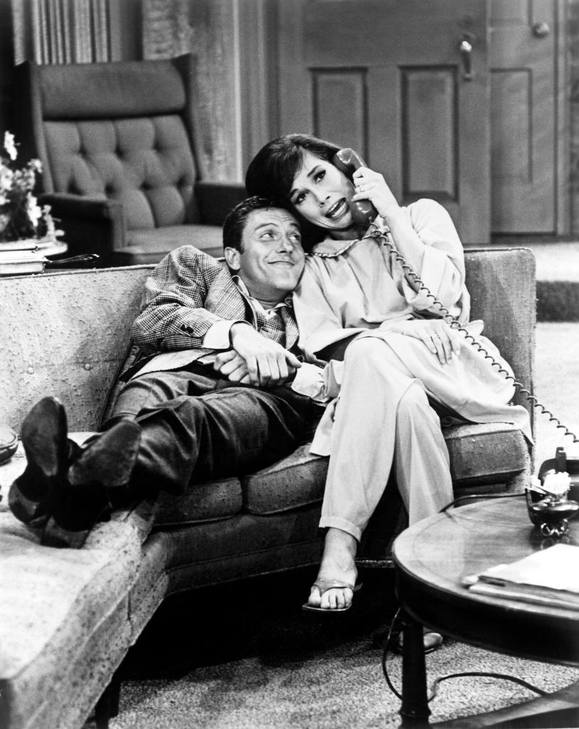 A black-and-white photo of Dick Van Dyke and Mary Tyler Moore on the living-room set of "The Dick Van Dyke Show," which aired on CBS from 1961-1966. They're seated on the couch; Dick is lounging out and has his head on Mary's shoulder while she holds the telephone to her ear. The rotary telephone is on the table in front of them.