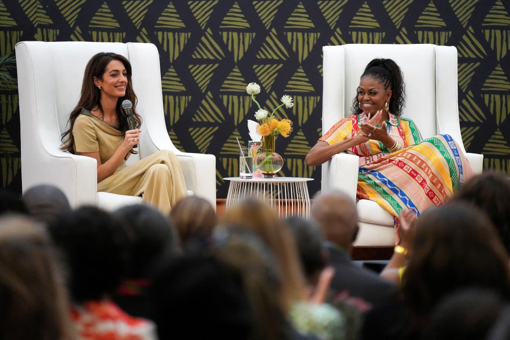 Michelle Obama smiling and clapping while Amal Clooney speaks into a microphone